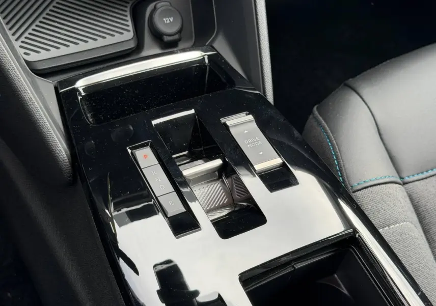 Gros plan sur la console centrale noire brillante avec levier de vitesses automatique et bouton Drive Mode, siège tissu gris visible.
