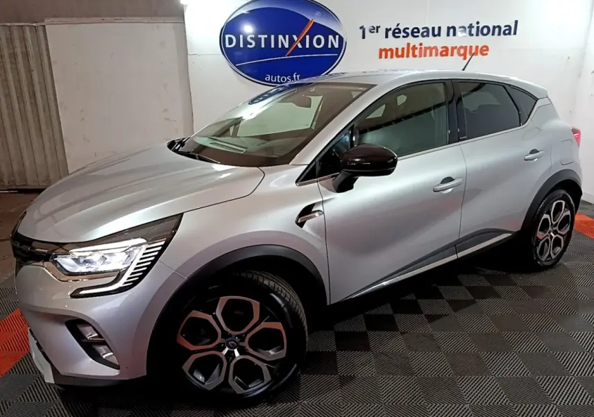 Renault Captur Intens E-Tech hybride rechargeable gris clair, vue 3/4 avant droit avec jantes distinctives et feux LED allumés.