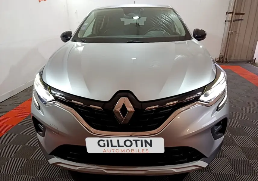 Vue avant d'un Renault Captur Intens E-Tech hybride rechargeable gris clair avec phares LED allumés en intérieur showroom.