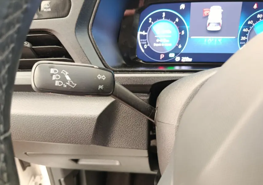 Gros plan sur le commodo gauche du volant d’un Volkswagen Caddy 2.0 TDI blanc, avec tableau de bord numérique en arrière-plan.