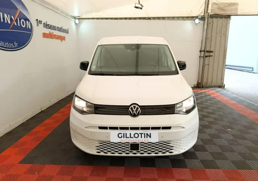Vue avant d'un Volkswagen Caddy blanc 2022 avec calandre noire et logo VW centré, en intérieur sur sol damier rouge et noir.