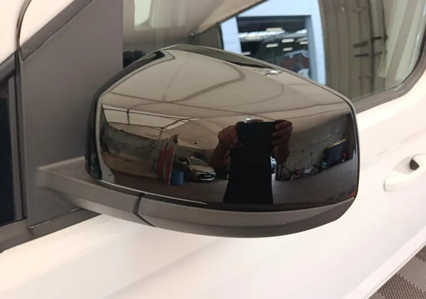 Rétroviseur extérieur droit noir brillant sur Volkswagen Caddy blanc, vue rapprochée en intérieur d'atelier.