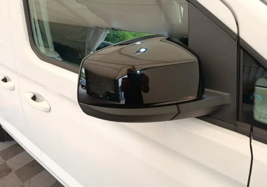 Rétroviseur noir côté conducteur d’un Volkswagen Caddy blanc vu de près, avec reflet de l’environnement intérieur.