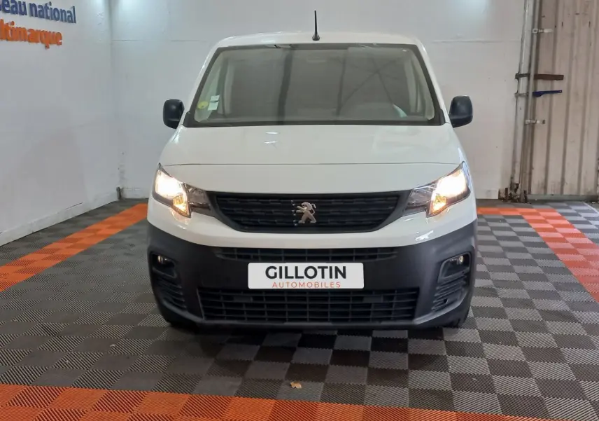 Vue avant d'un Peugeot Partner blanc 2021 avec phares allumés et pare-chocs noir dans un showroom.