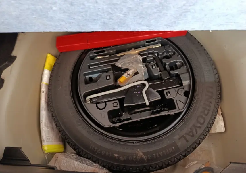 Vue plongeante sur la roue de secours noire et ses outils de bord dans le coffre d'une Peugeot 208 blanche.