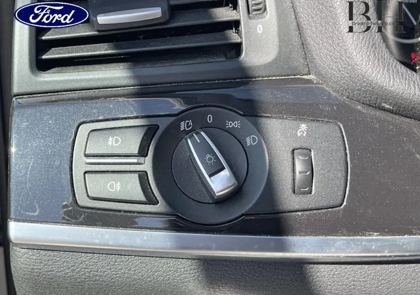 Gros plan sur la commande des phares d’un tableau de bord noir, avec logo Ford visible en haut à gauche.