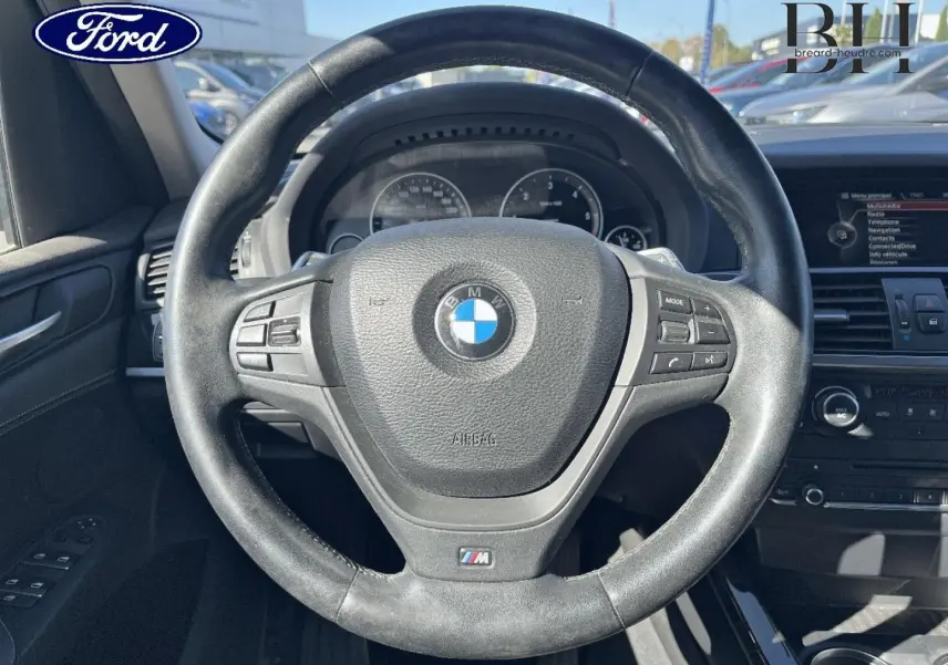 Vue rapprochée du volant cuir BMW M avec commandes intégrées, tableau de bord et écran multimédia visibles en arrière-plan.