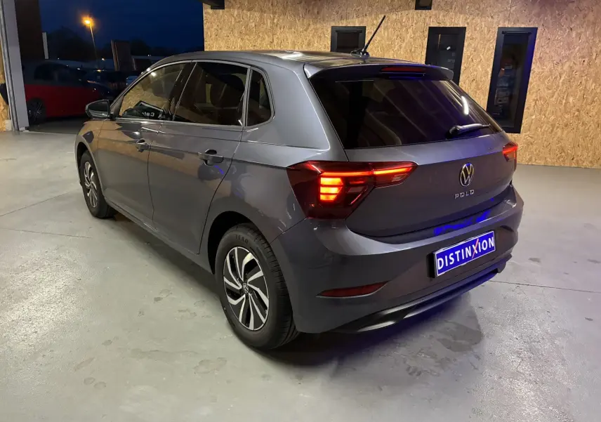 Volkswagen Polo gris vue 3/4 arrière droit en intérieur, feux arrière allumés et jantes bi-ton visibles.