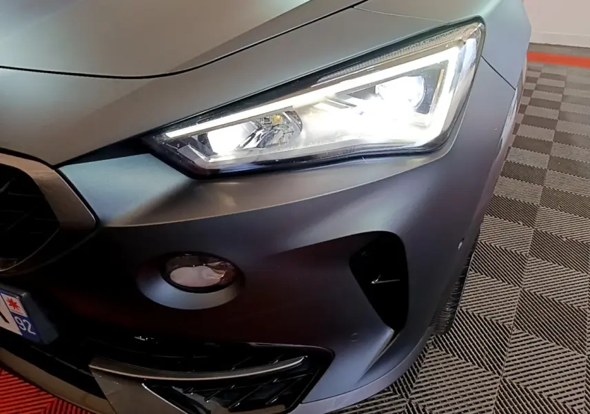 Gros plan sur l'avant droit d'une CUPRA Formentor gris foncé, mettant en valeur le phare LED allumé et la calandre sportive.