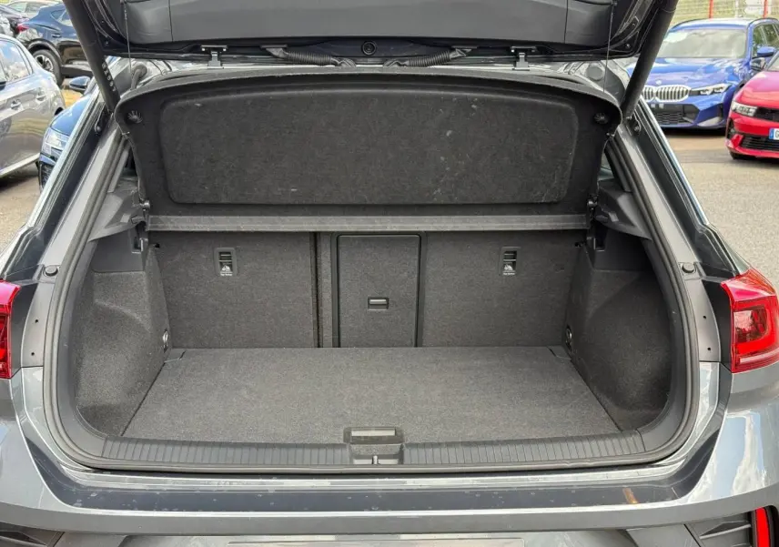 Coffre ouvert vu de l'arrière d'un Volkswagen T-Roc 2025 gris Indium métallisé avec intérieur de coffre spacieux et tapis gris.
