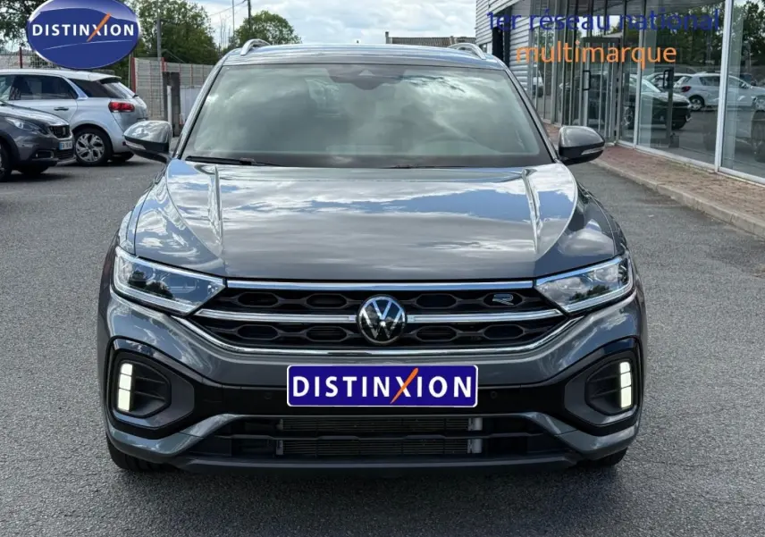 Vue avant d’un Volkswagen T-Roc gris Indium métallisé avec calandre R-Line et feux LED allumés sur un parking extérieur.
