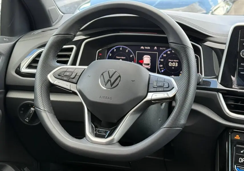 Vue rapprochée du volant multifonction noir et gris du Volkswagen T-Roc 2025 avec tableau de bord numérique en arrière-plan.