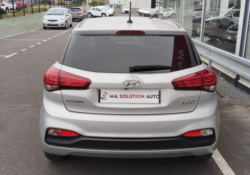 Vue arrière d'une Hyundai i20 gris argenté avec feux arrière rouges et vitre arrière teintée, stationnée en extérieur.