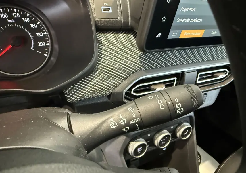 Gros plan intérieur sur la commande d’essuie-glace à gauche du volant du Dacia Sandero gris 2025, avec tableau de bord et écran tactile.
