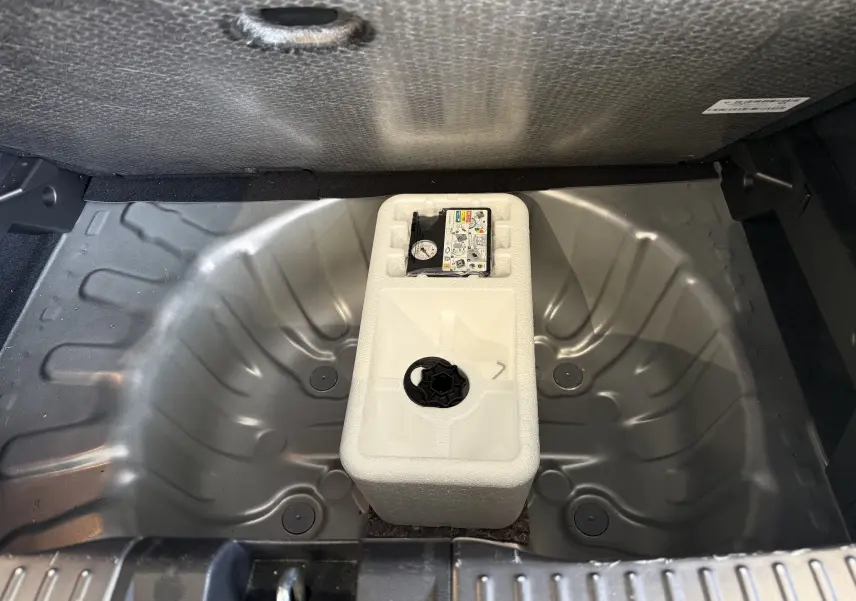 Vue plongeante sur le coffre nu gris d'une Dacia Sandero 2025, montrant le logement vide pour roue de secours avec kit de réparation.