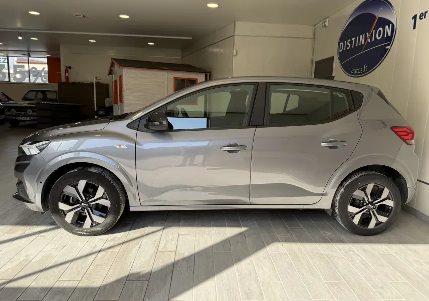 Vue de profil côté gauche d'une Dacia Sandero gris clair 2025 dans un showroom lumineux avec jantes noires et argentées.