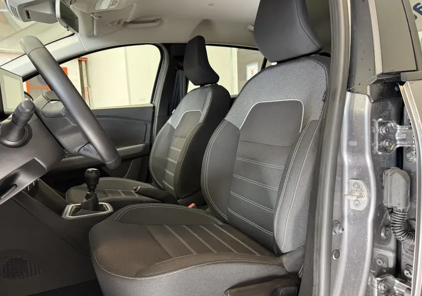 Vue intérieure côté conducteur de la Dacia Sandero gris 2025, sièges tissu noir avec surpiqûres blanches et boîte manuelle.