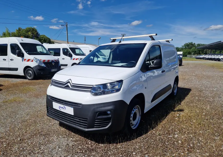 Vue 3/4 avant droite d’un Citroën Berlingo blanc 2020 avec barres de toit, garé sur un terrain ensoleillé.