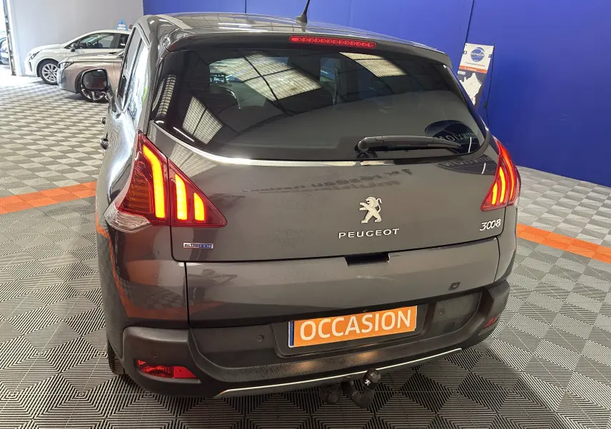 Vue arrière droite d’un Peugeot 3008 gris avec feux allumés, attelage visible et plaque orange « OCCASION » en intérieur showroom.