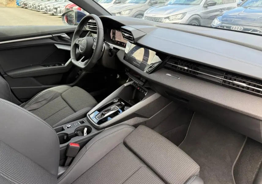 Vue intérieure côté conducteur de l'Audi A3 Sportback 2025, avec sièges tissu noir et tableau de bord moderne.
