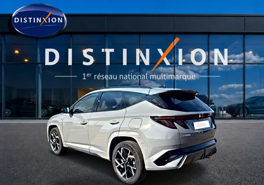 Vue 3/4 arrière gauche du Hyundai Tucson 2026 gris shadow avec jantes alu noires et toit panoramique en verre devant une vitrine Distinxion.