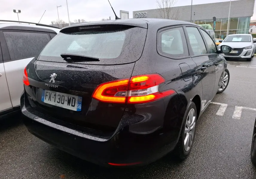 Peugeot 308 SW noire vue en 3/4 arrière droit sous la pluie avec feux arrière allumés sur parking.