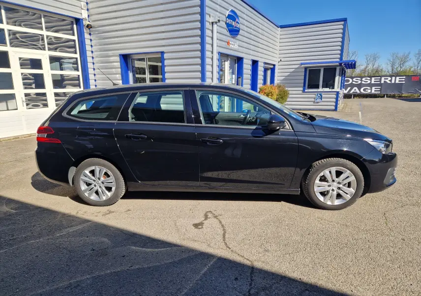 Peugeot 308 SW noire vue de profil côté gauche, stationnée devant un bâtiment industriel gris et bleu.
