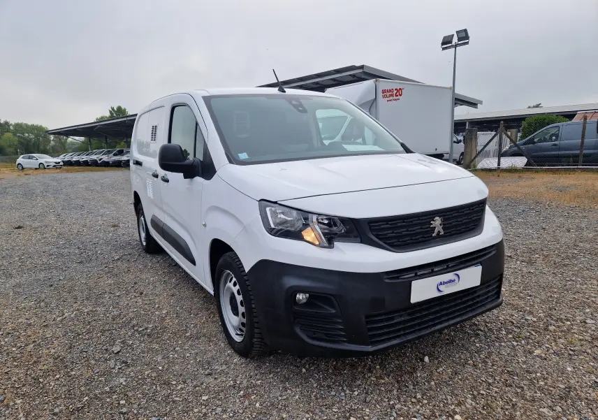 Peugeot Partner blanc vue 3/4 avant droit sur parking, avec calandre noire et feux allumés.