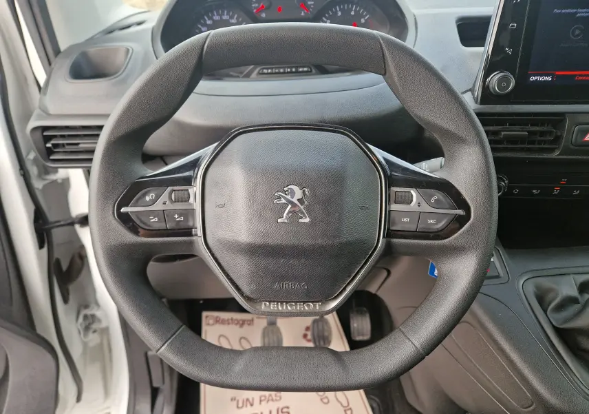 Vue rapprochée du volant noir du Peugeot Partner blanc 2021, avec commandes intégrées et tableau de bord en arrière-plan.