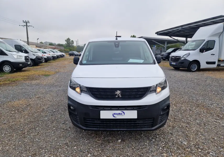 Vue frontale d'un Peugeot Partner blanc 2021 avec calandre noire et phares allumés sur un parking de véhicules utilitaires.