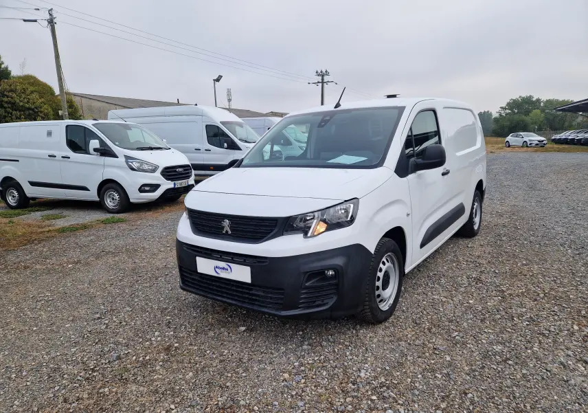 Peugeot Partner blanc en 3/4 avant droit sur un sol gravillonné, avec calandre noire et phares allumés.