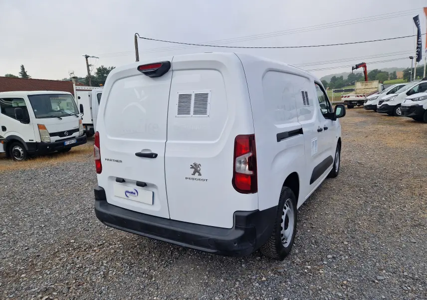 Vue 3/4 arrière droite d’un Peugeot Partner blanc utilitaire avec ventilation latérale sur un terrain gravillonné.