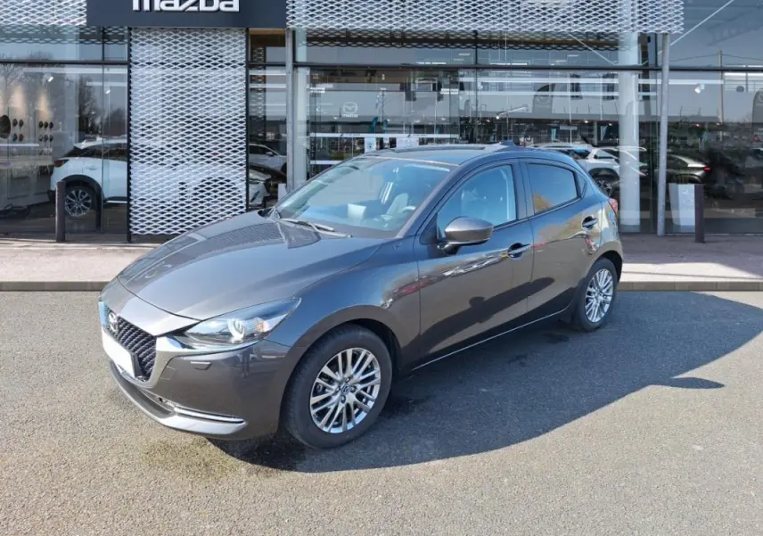 Vue 3/4 avant droit d'une Mazda2 gris métallisé garée devant un showroom Mazda avec jantes alliage.
