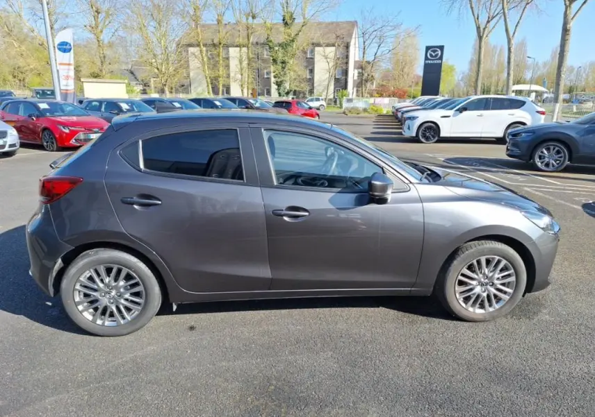 Profil côté gauche d'une Mazda2 gris métallisé 2023 avec jantes alliage et vitres surteintées sur parking extérieur.