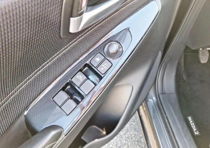 Vue rapprochée du panneau de commandes des vitres côté conducteur d'une Mazda2 gris 2023 avec boutons auto et réglage rétroviseurs.