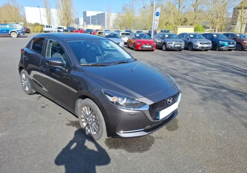 Vue 3/4 avant gauche d'une Mazda2 gris métallisé avec jantes aluminium sur un parking extérieur.