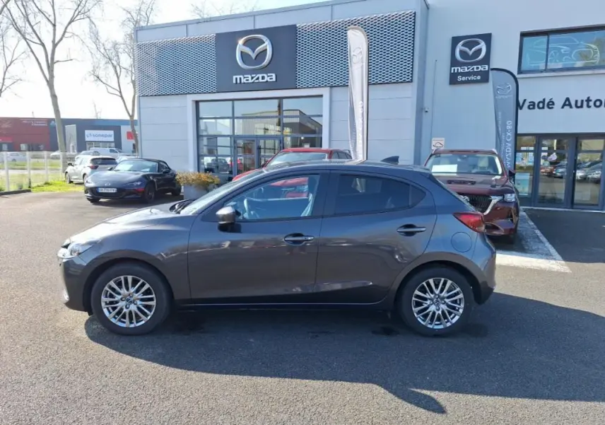 Vue de profil côté gauche d'une Mazda2 gris métallisé stationnée devant un concessionnaire Mazda.