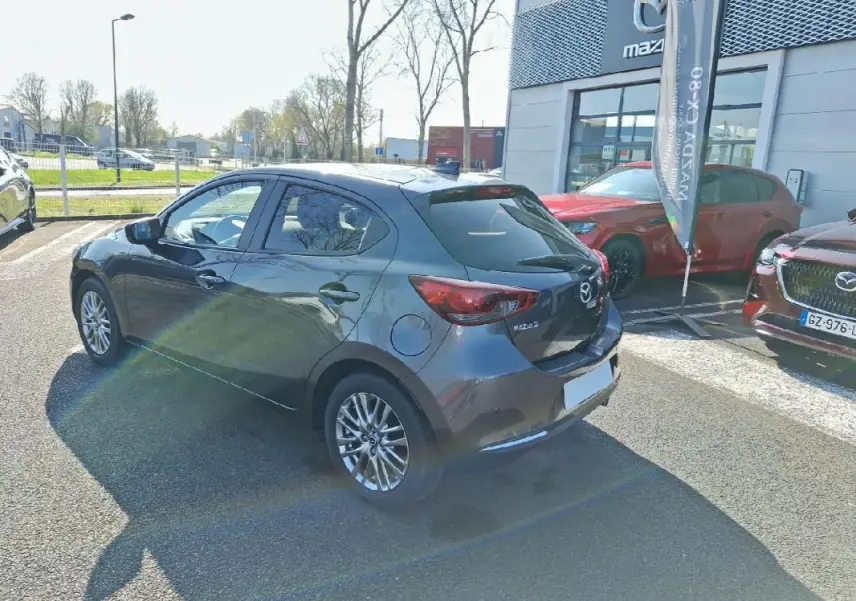 Vue 3/4 arrière droite d'une Mazda2 gris métallisé 2023 avec jantes aluminium, stationnée devant un concessionnaire Mazda.