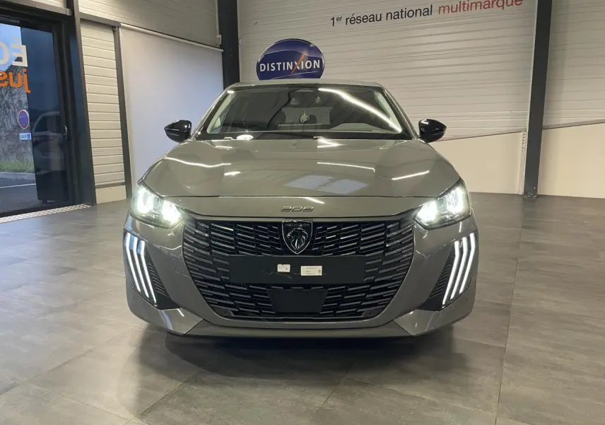 Vue avant d'une Peugeot 208 gris selenium avec phares allumés et signature lumineuse verticale LED visible en intérieur showroom.