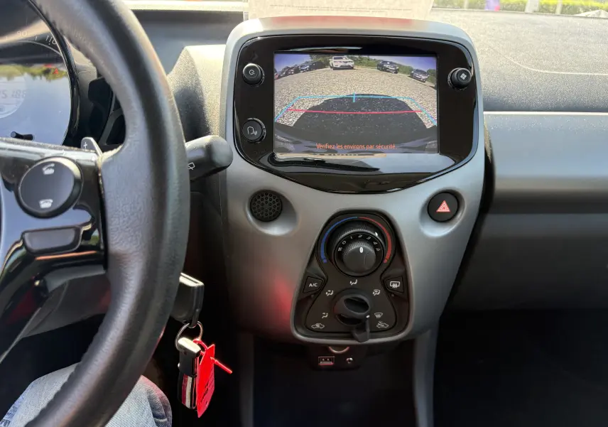 Vue intérieure centrée sur la console centrale d'une Toyota Aygo MC blanche, écran de caméra de recul et commandes climatisation visibles.