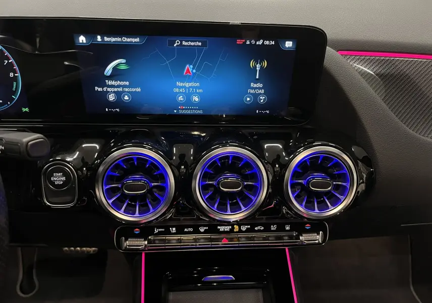 Vue intérieure du tableau de bord de la Mercedes GLA 250E AMG Line 2023, avec aérateurs ronds éclairés en bleu et ambiance lumineuse rose.