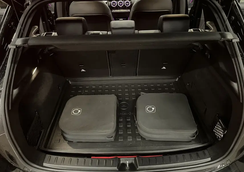 Coffre ouvert d’un Mercedes GLA 250E AMG Line noir Cosmos, avec tapis de coffre caoutchouc et deux housses noires.
