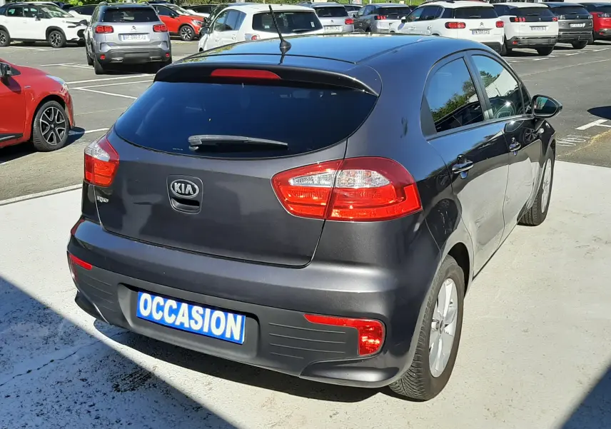Vue 3/4 arrière droite d'une KIA RIO grise 5 portes de 2016 avec feux arrière allumés sur parking extérieur.