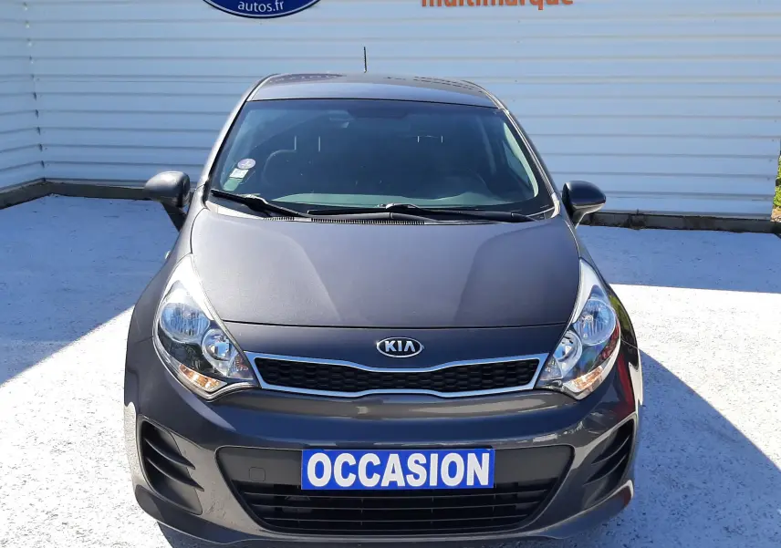 Vue frontale d'une KIA Rio grise 2016 avec plaque "OCCASION" et calandre caractéristique, stationnée devant un mur blanc.