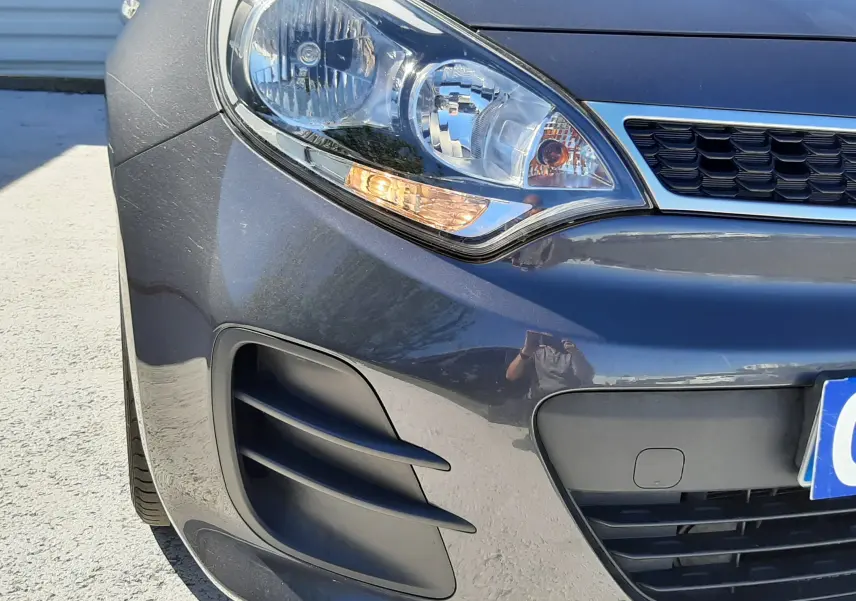 Gros plan sur l'avant droit d'une KIA Rio grise 2016, mettant en valeur le phare et la calandre noire.