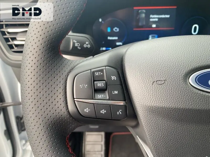 Gros plan sur le volant perforé noir avec commandes multifonctions du Ford Kuga blanc Glacier, tableau de bord digital en arrière-plan.