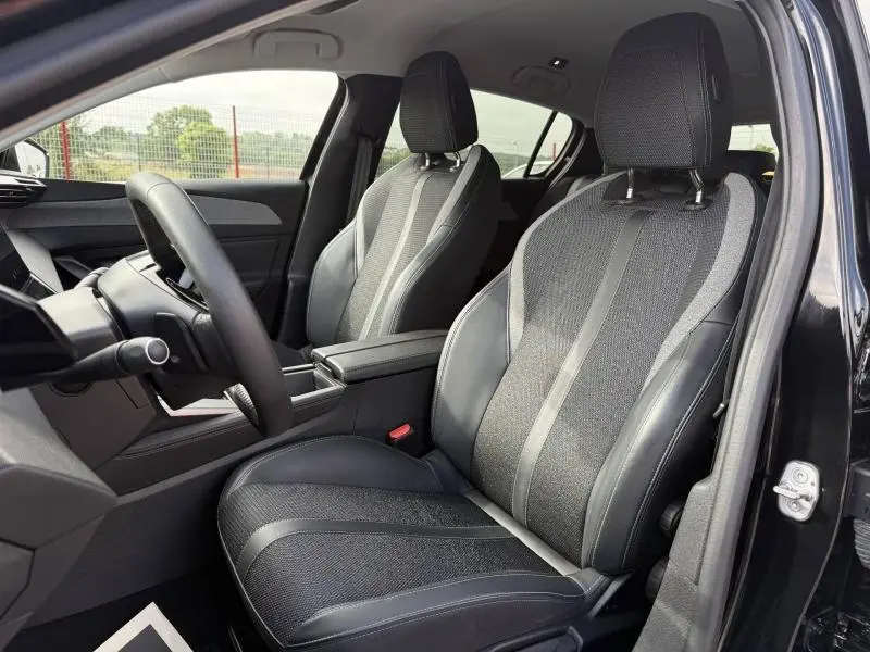 Vue intérieure côté conducteur de la Peugeot 308 2024 noir Perla Nera, sièges en tissu et cuir avec accoudoir central.