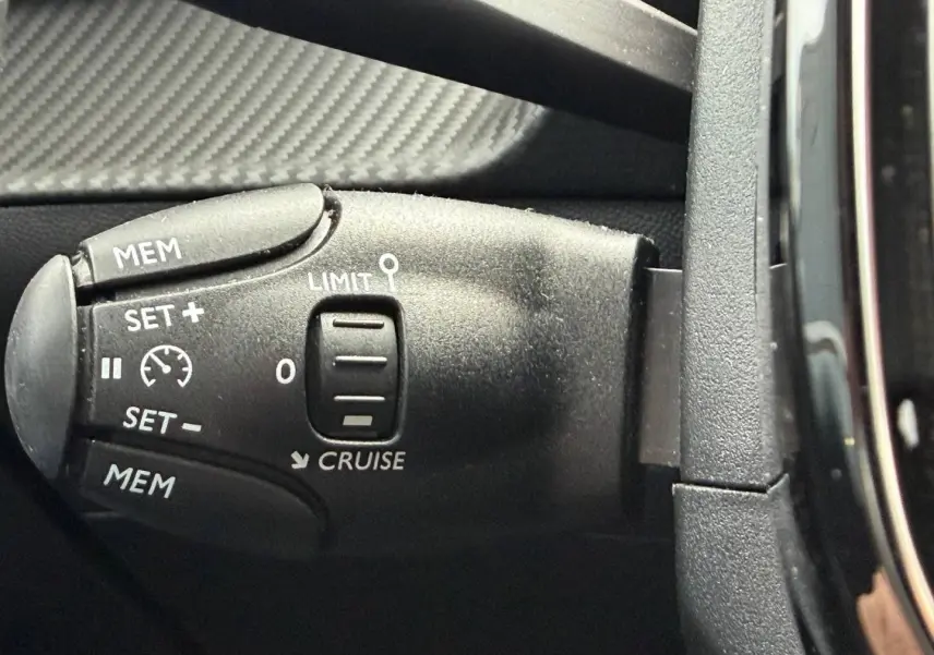 Gros plan sur la commande de régulateur de vitesse noire à gauche du volant dans une Peugeot 208 noire Perla Nera 2025.
