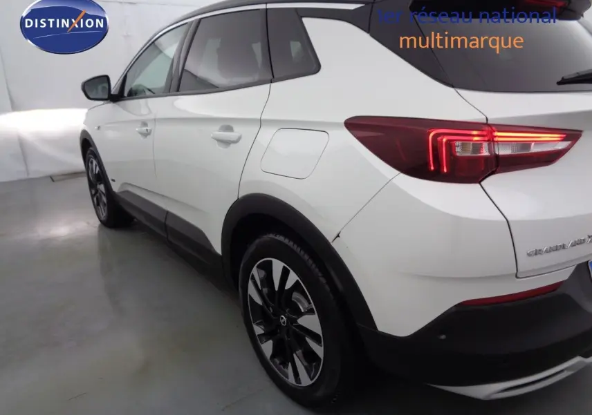 Vue 3/4 arrière côté gauche d'un Opel Grandland X blanc perle métallisé avec feux arrière allumés et jantes noires bicolores.