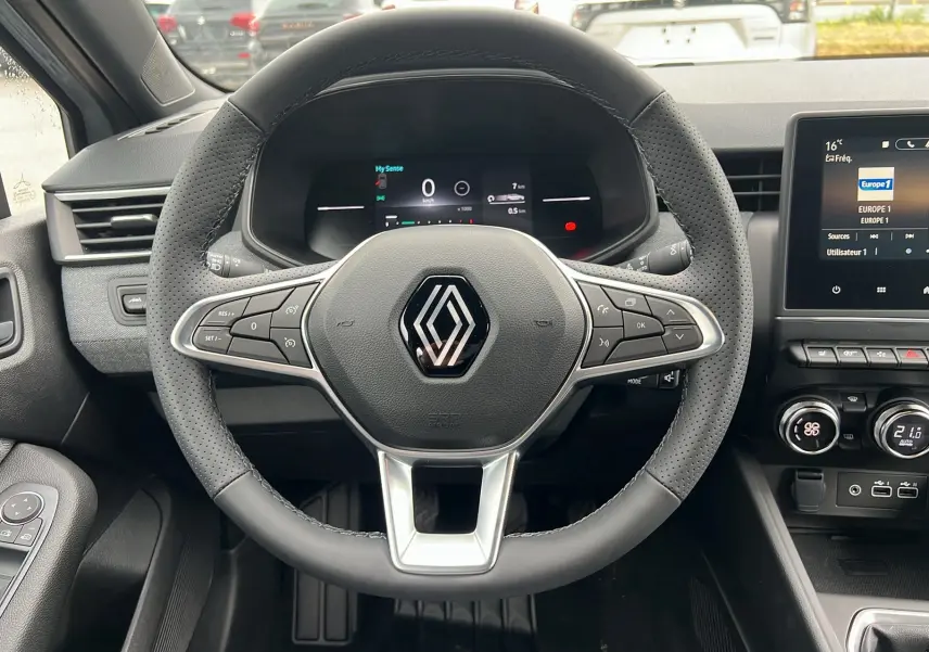 Vue intérieure centrée sur le volant perforé de la Renault Clio V TCE 90 Techno 2025 avec tableau de bord digital et écran tactile.
